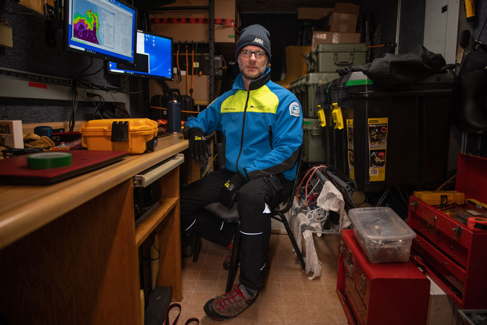 Matthew Shupe, wearing an ARM beanie, blue-and-yellow jacket, and black pants, swivels to face the camera in his container office in the bow of the Polarstern.