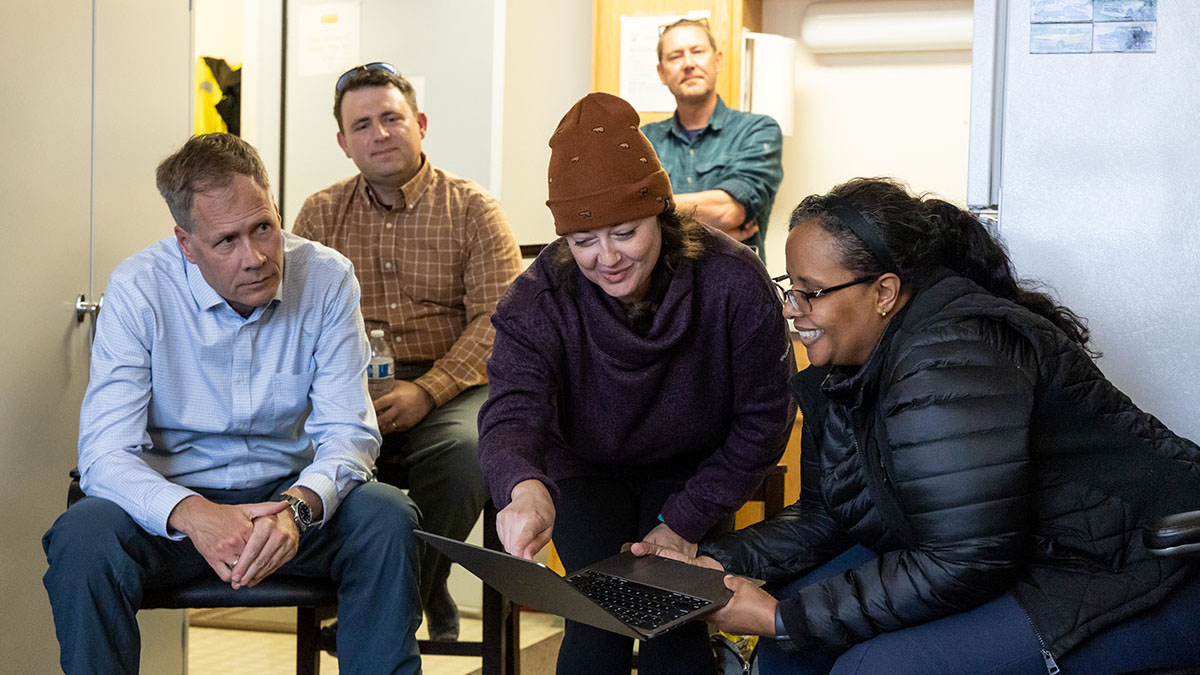 A person leans over to point something out on a laptop screen to Dr. Berhe, who is sitting down and smiling while holding the bottom of the computer.