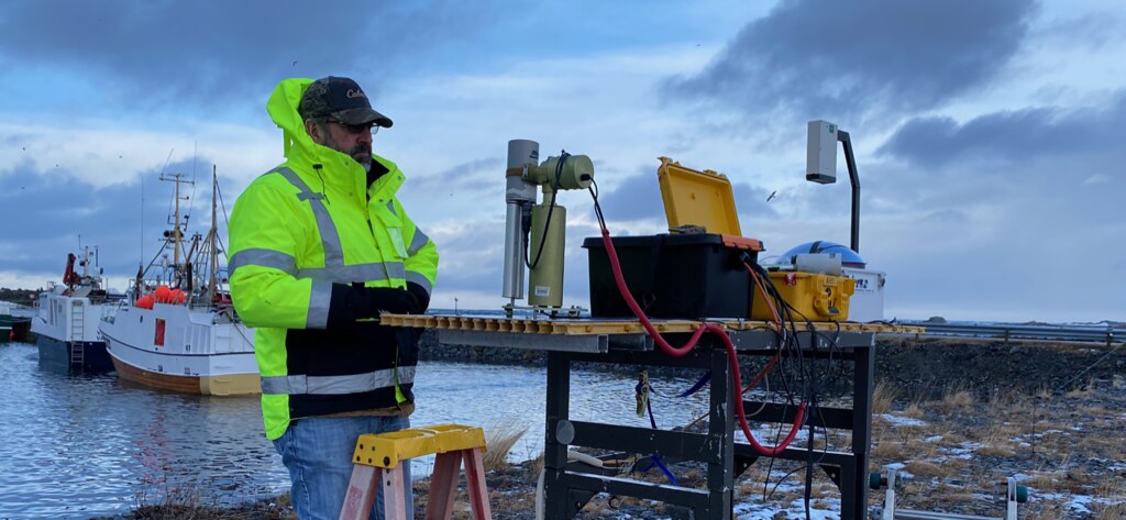 Wearing a bright yellow safety jacket, John Hamelmann looks at a sun photometer on a worktable. A boat is on the water behind Hamelmann. 