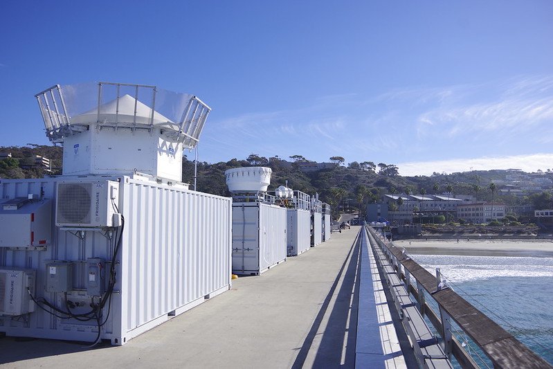 Five ARM containers with instruments on top line Scripps Pier. Buildings and trees near the shore are visible.