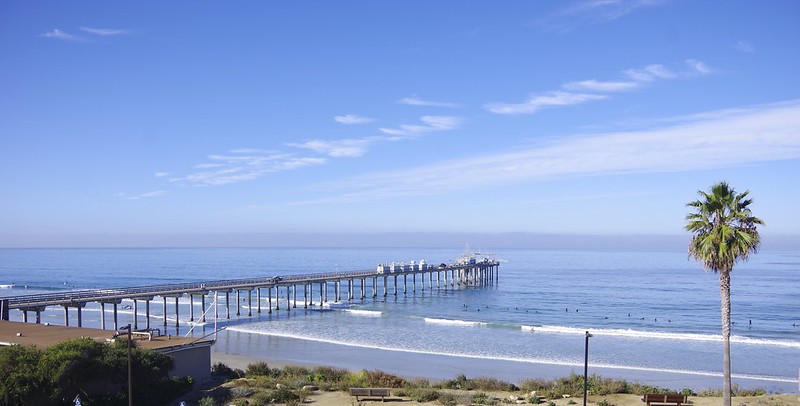 With clouds overhead and along the horizon, the Scripps Pier juts out into the Pacific Ocean. ARM instruments and containers are visible in the middle of the pier. To the right of the pier, people play in the water. A palm tree stands at the far right of the picture, with a bench and a building closer to the pier.
