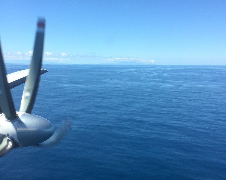 The picture shows blue sky with deck-like clouds in the distance and calm blue ocean. The camera captures a plane propeller spinning to the left.