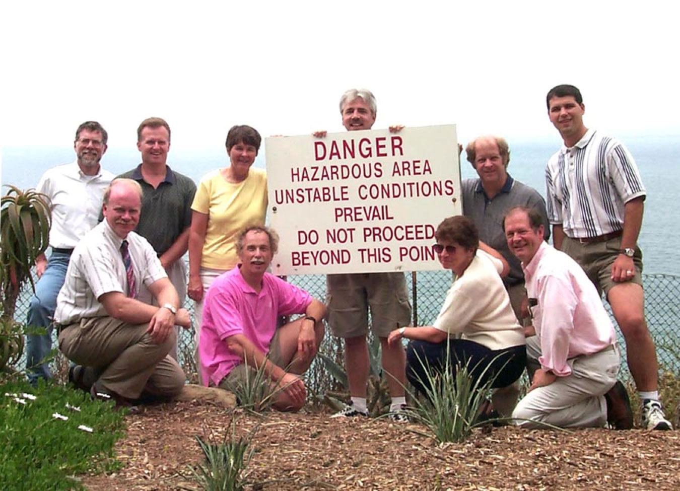 Ten ARM Infrastructure Review Committee members stand with a sign, held by Kevin Widener, that says, "Danger, Hazardous Area, Unstable Conditions Prevail, Do Not Proceed Beyond This Point." The photo was taken July 14, 1999, in La Jolla, California, overlooking the Pacific Ocean.