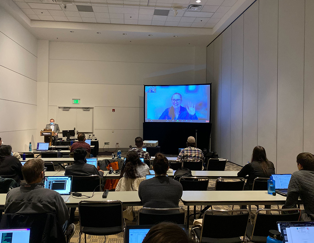 Students are seated at tables with laptops open, and Max Grover stands at a podium next to a big-screen TV showing Allison Aiken waving to the class.