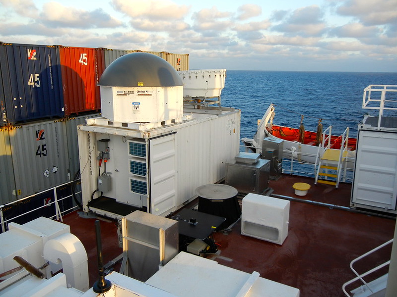 ARM radars are surrounded by cargo containers on a ship traveling in the Pacific Ocean. Puffy white clouds are visible overhead.