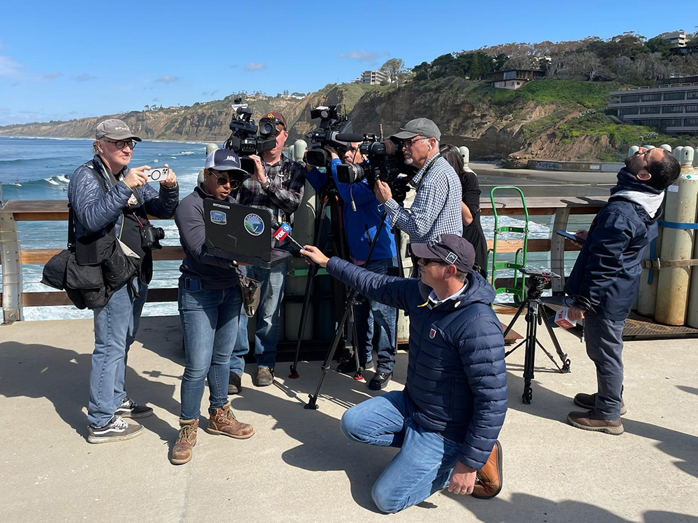 On Scripps Pier, journalists with cameras and microphones crowd around Gabi Pessoa as she points out weather balloon data appearing on a laptop screen.