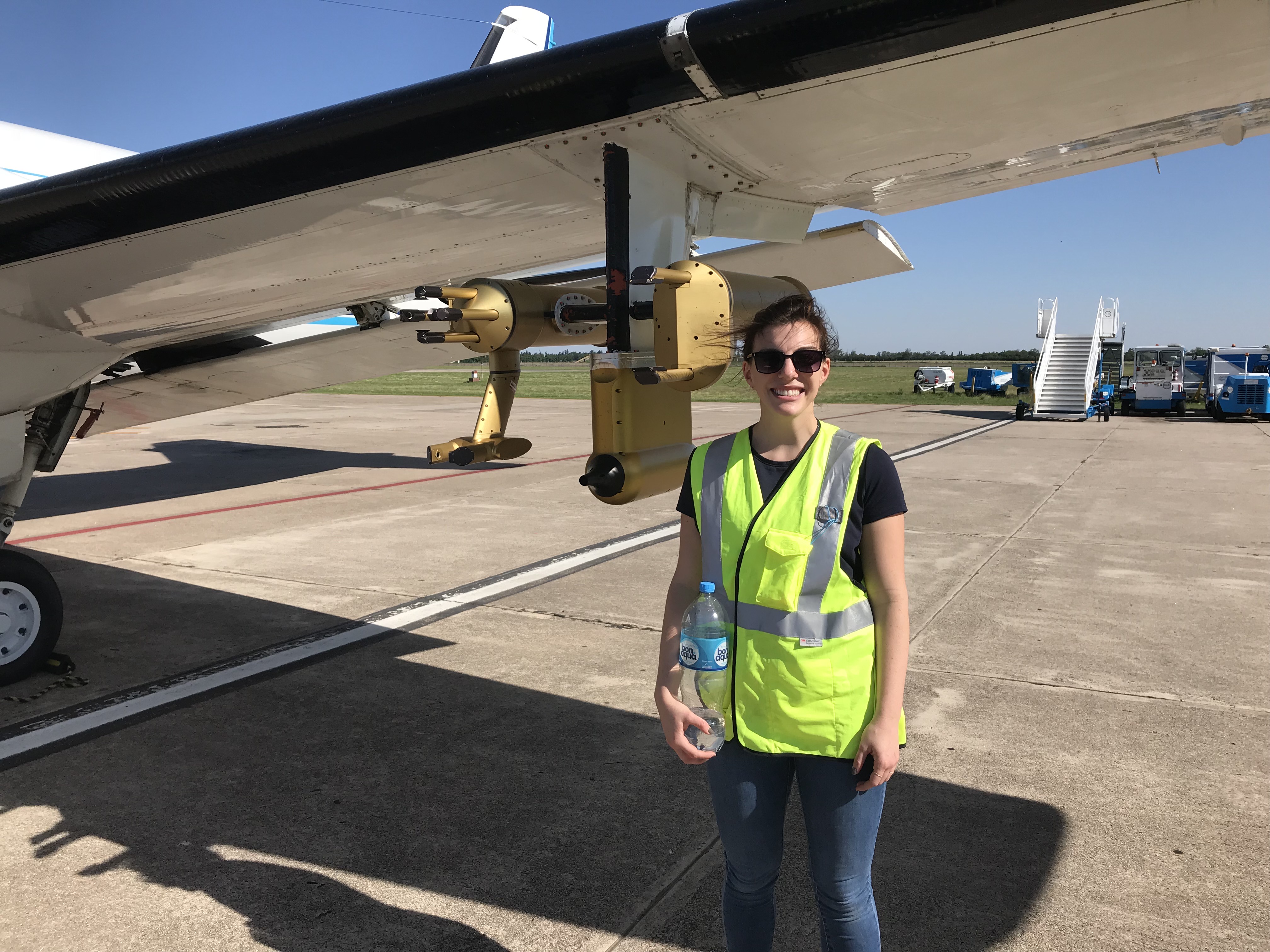 Wearing sunglasses, a bright yellow safety vest, and jeans, Lexie Goldberger stands next to an aircraft.