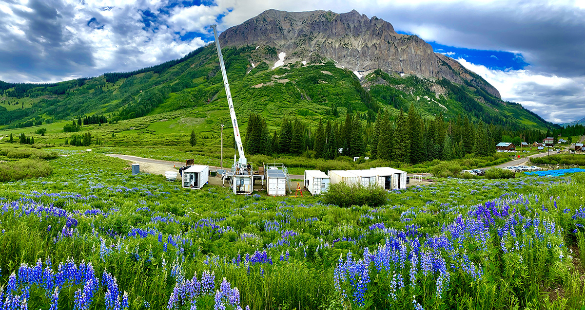 Eight cargo containers are in place at Gothic Townsite, Colorado, in front of Gothic Mountain, which juts into a vibrant blue sky with clouds. A field of lupine blooms in the foreground.