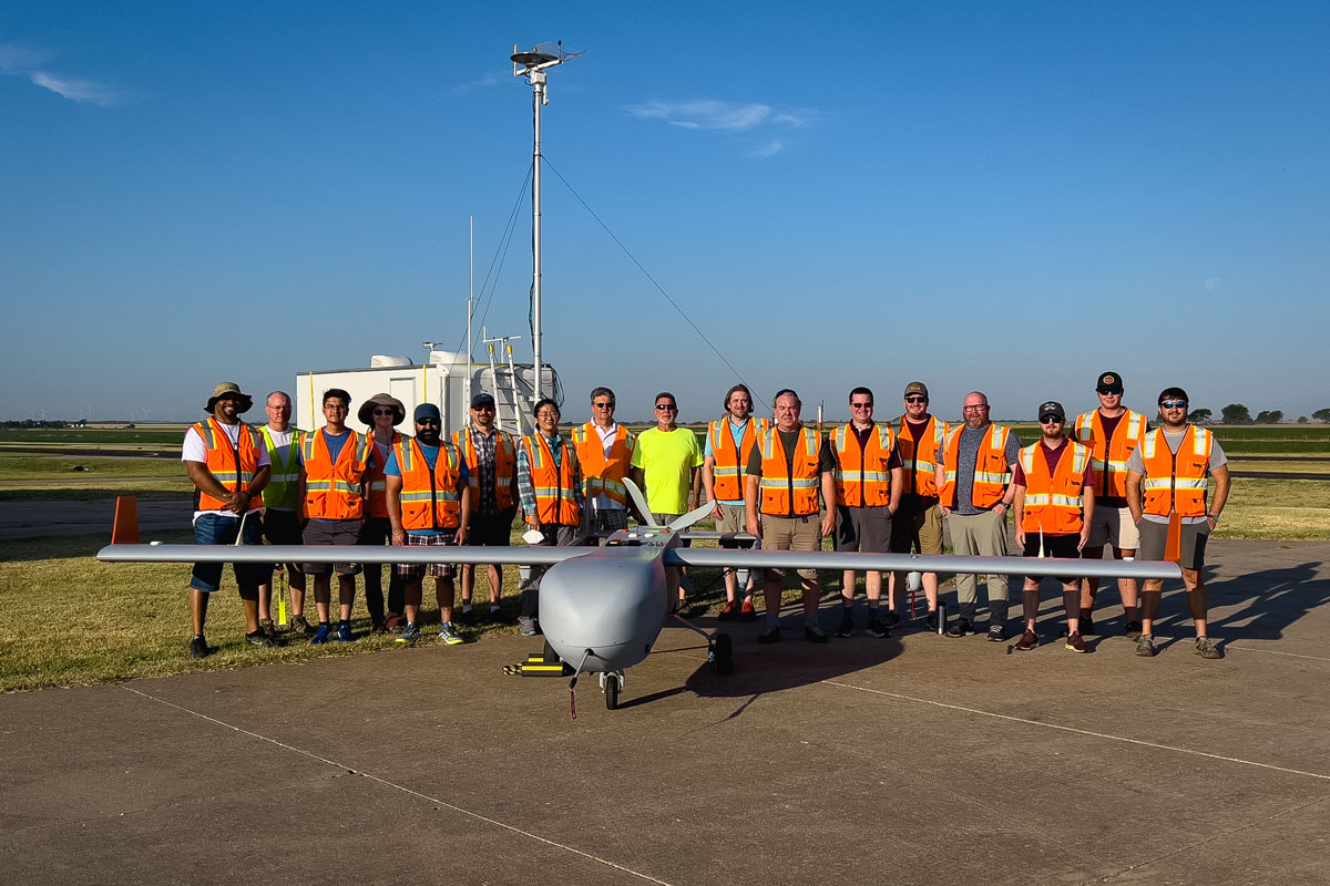 Seventeen ARM team members--all but one wearing orange safety vests and the other wearing a bright yellow T-shirt--stand behind the ArcticShark uncrewed aerial system in Oklahoma.
