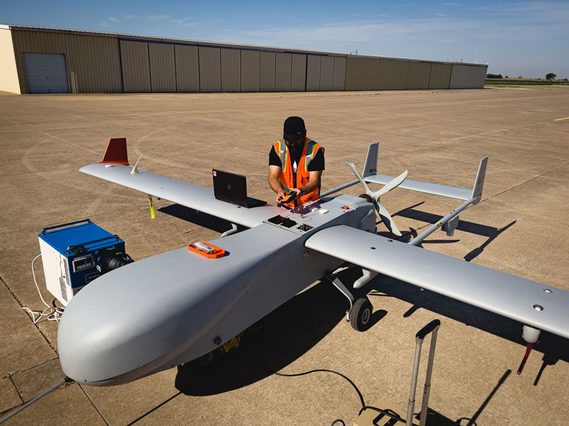 Hardeep Mehta kneels while working on the ArcticShark uncrewed aerial system, with an open laptop resting on its wing.