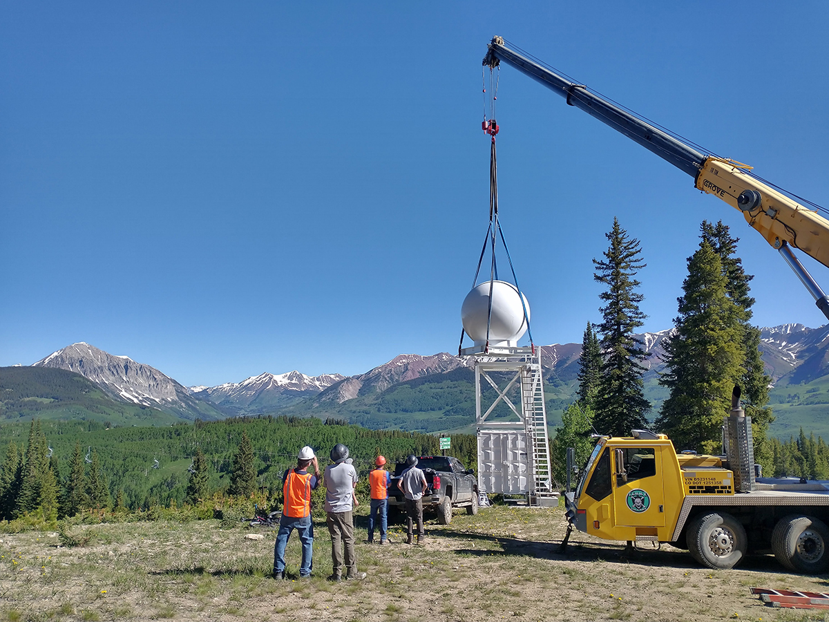 A crane lifts a white radar dome off its platform as technicians wearing hard hats watch from the ground.
