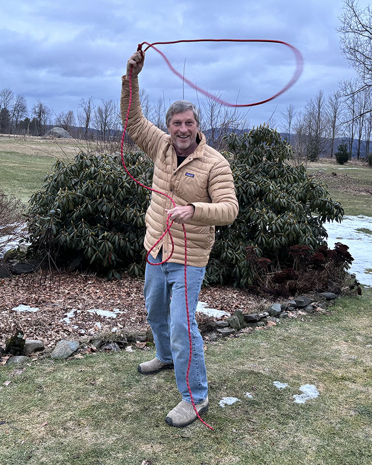 Andrew Vogelmann twirls a red lasso around his head while standing outside in front of a bush.