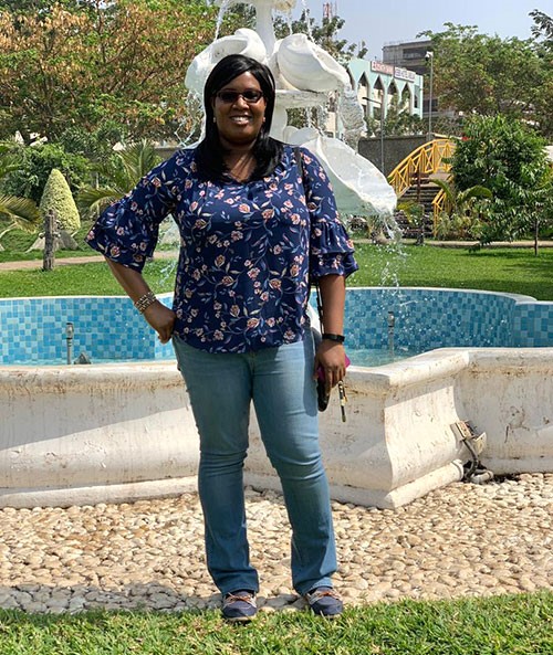 Ogo Enekwizu, wearing sunglasses, a blue blouse, and jeans, stands in front of a white stone fountain.