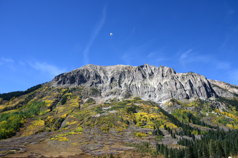 A white balloon hovers in the sky. Nearby, green and yellow dots the mountainside below.