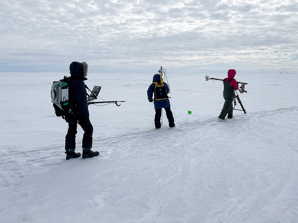 Under a cloudy sky, one person holds a computer while two others collect snow measurements on Alaska's Elson Lagoon.