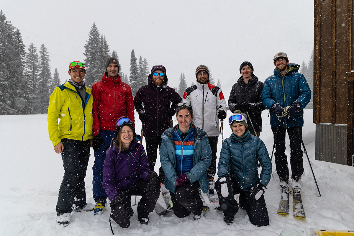 Nine people pause for a picture in falling snow. All are wearing winter jackets and pants. Some are on skis, and others are wearing snowboots.