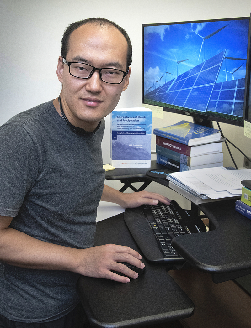 Yunpeng Shan turns away from his computer to look at the camera. A book on cloud and precipitation microphysics stands up on his desk near his left hand.