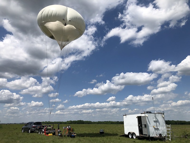 In a field next to a trailer, a tethered balloon system is winched into the sky.