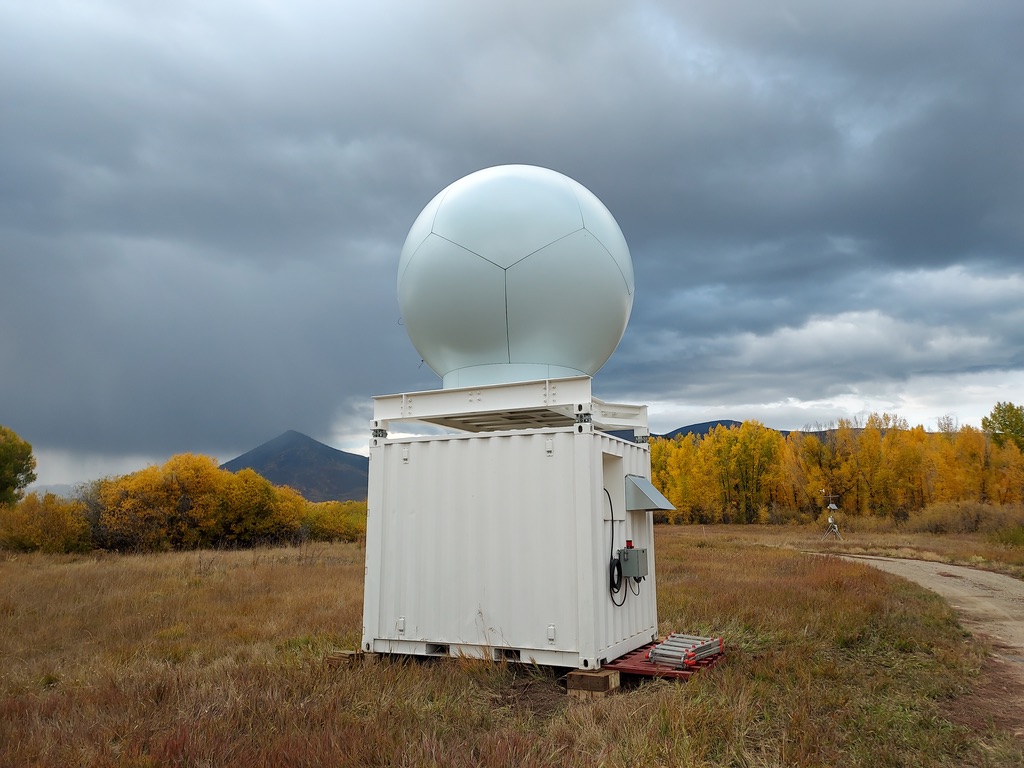 Under dark clouds, the X-band radar sits on top of a box in a grassy space. Nearby trees are changing color from green to yellow.