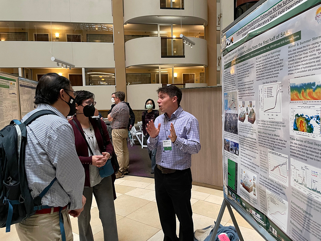 Daniel Feldman stands next to his poster and gestures while talking with Shaima Nasiri and Qiang Fu.