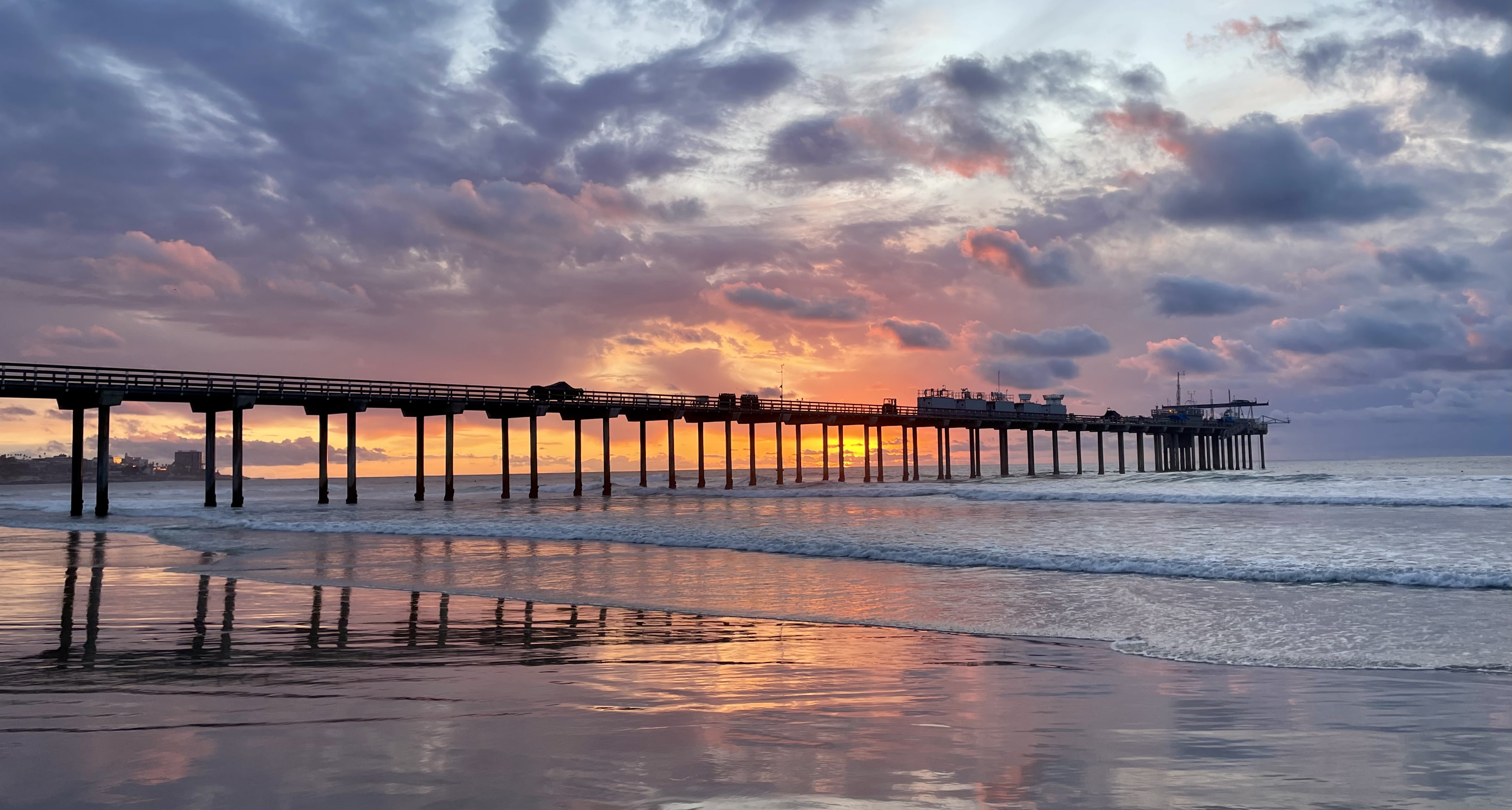 Waves wash ashore near the Ellen Browning Scripps Memorial Pier, which is hosting the ARM Mobile Facility for EPCAPE. The pier sits under a cloudy purple-orange sky.