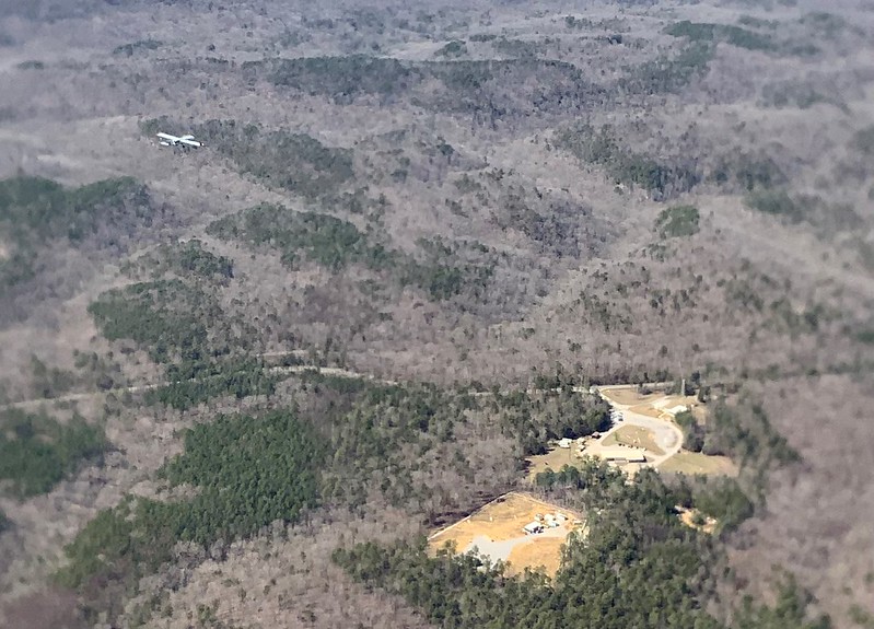 An uncrewed aerial system flies over a forest with buildings and roads visible in the middle of clearings. Patches of brown and green are seen throughout the forest.