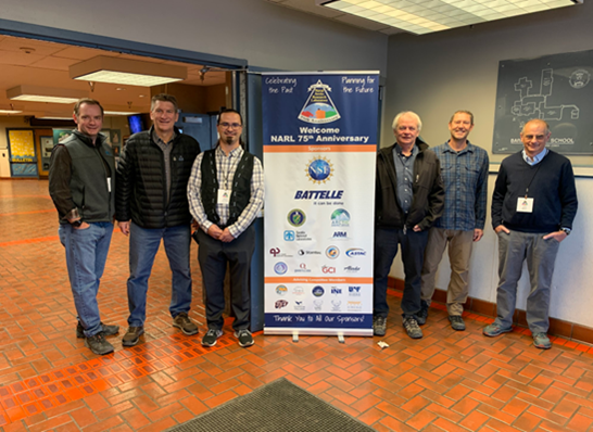 Six men stand in a hallway next to a sign that says "Welcome NARL 75th Anniversary."