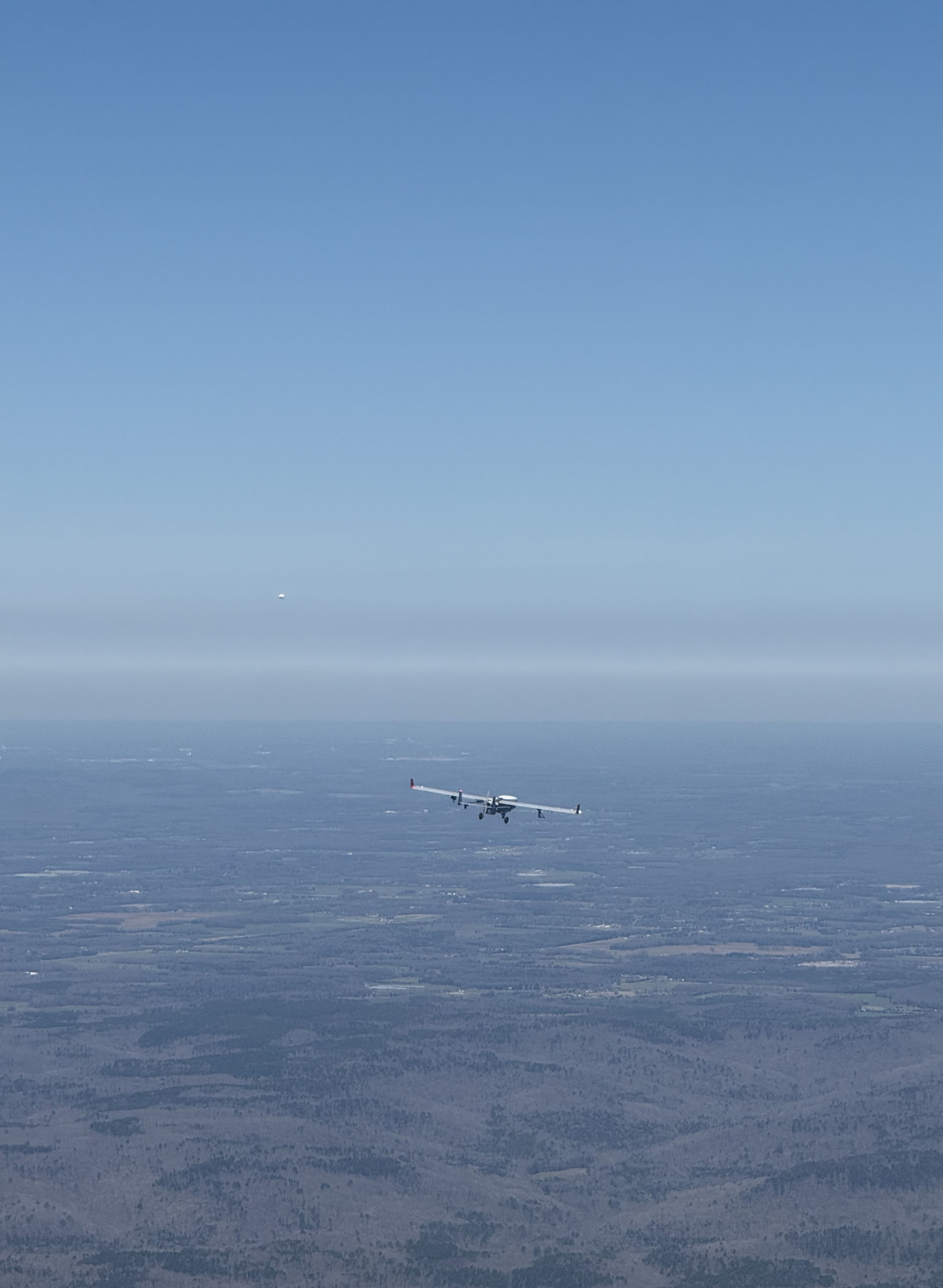 An uncrewed aerial system flies above a forest while a white dot is seen in the distance.