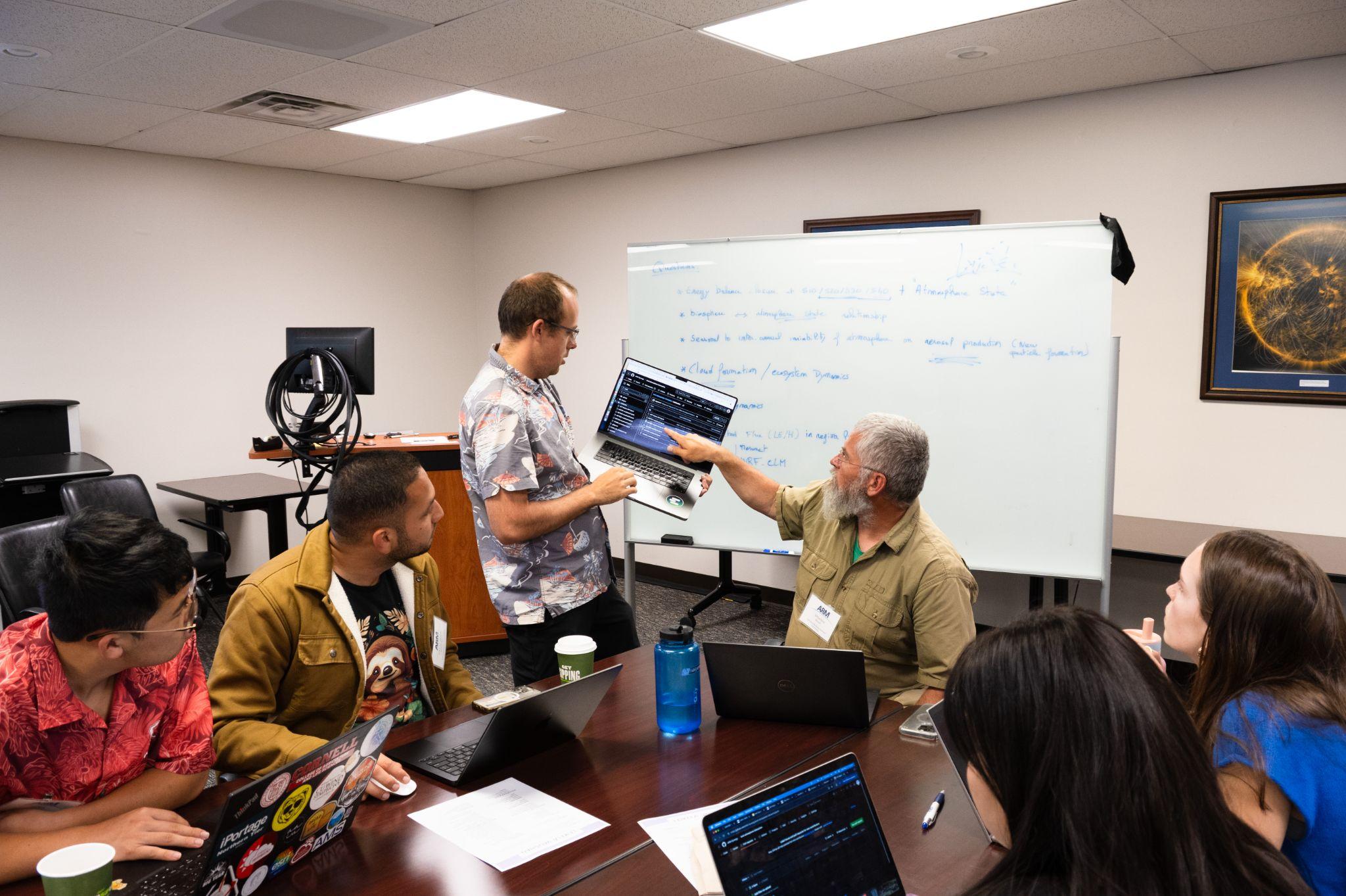 A person points to a laptop being held by another person while a group of students around a classroom table look on.