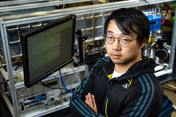 Zezhen “Jay” Cheng poses next to a computer screen and instrument rack with his arms folded.