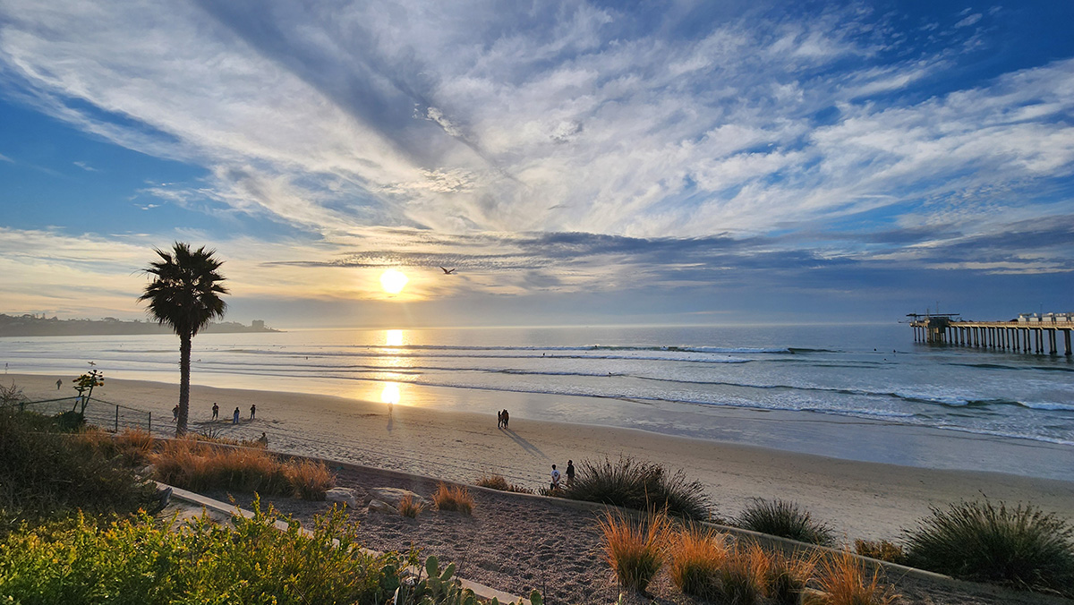 In this January 2024 photo, Scripps Pier, arrayed with ARM containers and instruments, juts out into the Pacific Ocean. Beachgoers walk along the water, and others talk while standing on the sand. The sun shines in the distance, illuminating clouds streaking across the sky and a piece of land downshore. A giant palm tree stands near the sand.